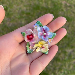 May include: A floral brooch featuring a cluster of colorful flowers. The brooch includes a pink rose, blue and purple pansies, and a yellow pansy, all with green leaves. The flowers are arranged in a star shape.