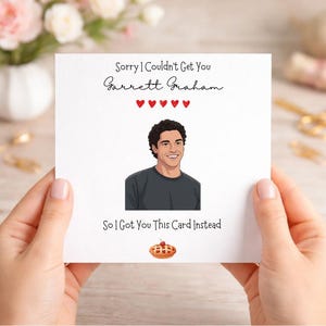 以下が含まれることがあります： 「Sorry I Couldn't Get You Garrett Graham」と「So I Got You This Card Instead」の文字が入った白いグリーティングカード。笑顔の人物と、さくらんぼが乗ったパイのイラストが描かれています。