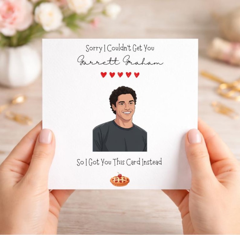 以下が含まれることがあります： 「Sorry I Couldn't Get You Garrett Graham」と「So I Got You This Card Instead」の文字が入った白いグリーティングカード。笑顔の人物と、さくらんぼが乗ったパイのイラストが描かれています。