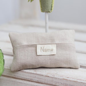 Könnte beinhalten: Ein kleines, beiges Leinenkissen mit einem weißen Etikett, auf dem "Name" steht. Das Kissen liegt auf einer weißen Holzoberfläche.