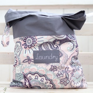 Könnte beinhalten: Graue und rosafarbene Wäschetasche mit Paisley-Muster und dem aufgenähten Wort "Laundry" auf der Vorderseite.