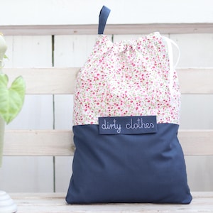 Könnte beinhalten: Ein weißer Wäschebeutel mit Kordelzug, floralem Muster und einem dunkelblauen Boden. Der Beutel hat ein Etikett mit der Aufschrift "dirty clothes".