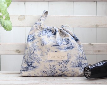May include: A white and blue toile patterned drawstring bag with a label that says "Hair dryer". A black hair dryer is sitting on a white wooden surface next to the bag.