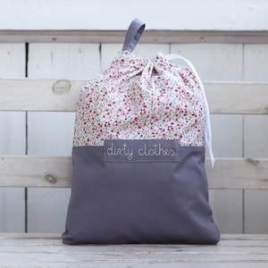 Könnte beinhalten: Eine graue und weiße Wäschetasche mit einem Blumenmuster und dem aufgenähten Text "dirty clothes" auf der Vorderseite.