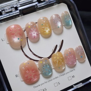 Peut inclure: Un ensemble de dix faux ongles dans un boîtier en plastique transparent, avec une base translucide ornée de motifs d'étoiles et de feux d'artifice colorés en rose, jaune et bleu. La carte présente le texte L:, R:, Thank you for your order, I hope you like it., XS, S, et M.