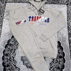 May include: A light gray tracksuit set featuring a hooded sweatshirt and matching sweatpants. The sweatshirt and sweatpants have the word "TRAPSTAR" embroidered in red, white, and blue. The set includes a drawstring and a tag.