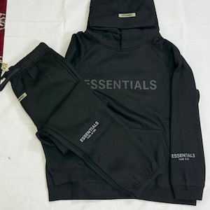 Peut inclure: Ensemble de survêtement noir composé d'un sweat à capuche et d'un pantalon de survêtement assorti. Le sweat-shirt porte le mot "ESSENTIALS" imprimé sur la poitrine et sur la manche. Le pantalon de survêtement porte également le mot "ESSENTIALS". L'ensemble est en matière souple.