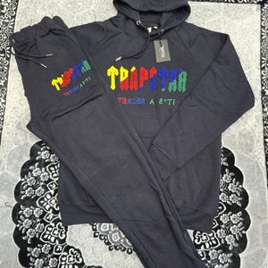 May include: A dark gray tracksuit set, including a hooded sweatshirt and matching sweatpants. The word "TRAPSTAR" is embroidered in colorful letters on both pieces. The set is displayed on a patterned surface.