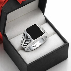 Peut inclure: Bague en argent avec une pierre centrale carrée noire, présentée dans un coffret cadeau noir avec un ruban rouge. La bague présente un motif de feuilles sur les côtés et le chiffre "925" est visible.