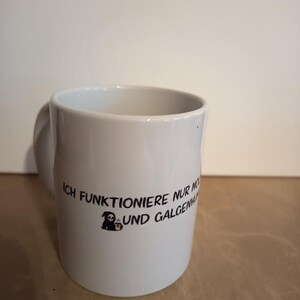 Op de afbeelding: Een witte keramische mok met zwarte tekst die luidt ICH FUNKTIONIERE NUR NOCH MIT KOFFEIN UND GALGENHUMOR, vergezeld van een kleine afbeelding van een grimmige maaier die een koffiekopje vasthoudt, aan de voorzijde van de mok.