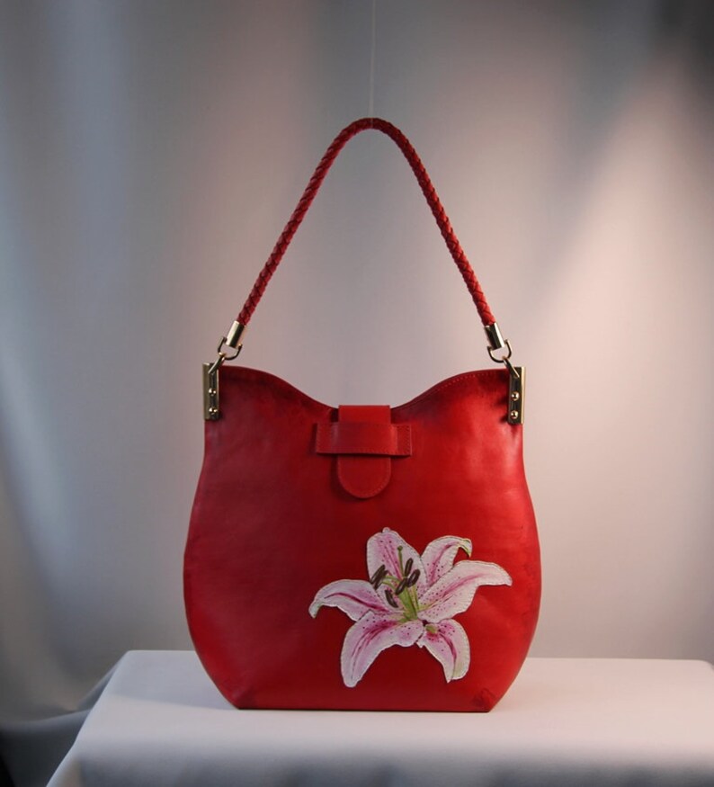 May include: A red leather handbag with a braided strap and a painted lily flower design.