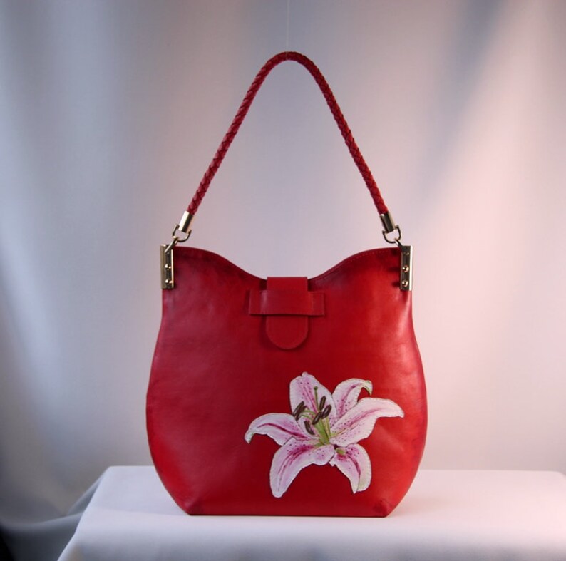 May include: A red leather handbag with a braided strap and a painted lily flower design.