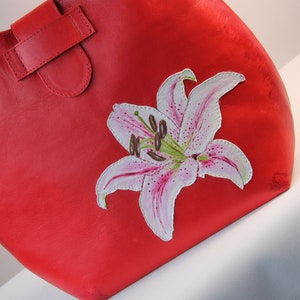 May include: A red leather tote bag with a hand-painted white lily flower design.