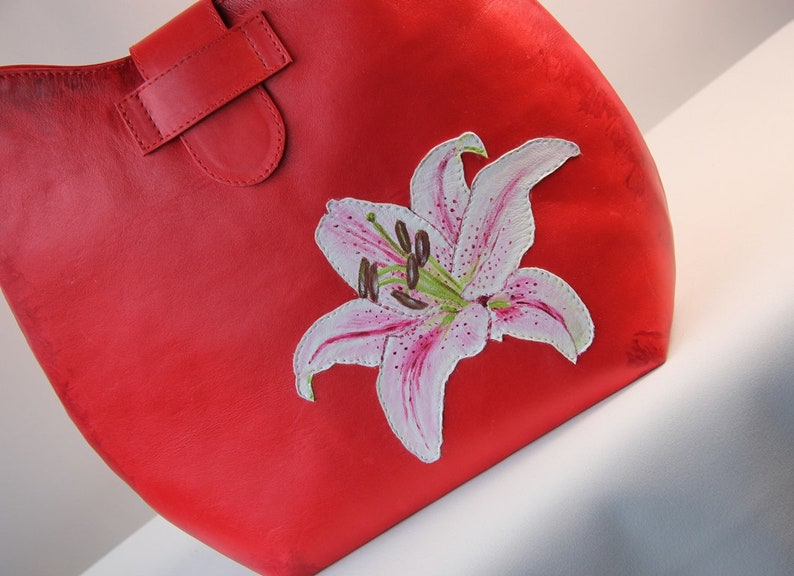 May include: A red leather tote bag with a hand-painted white lily flower design.