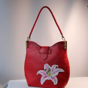 May include: Red leather handbag with a white lily flower painted on the front. The bag has a braided leather strap.