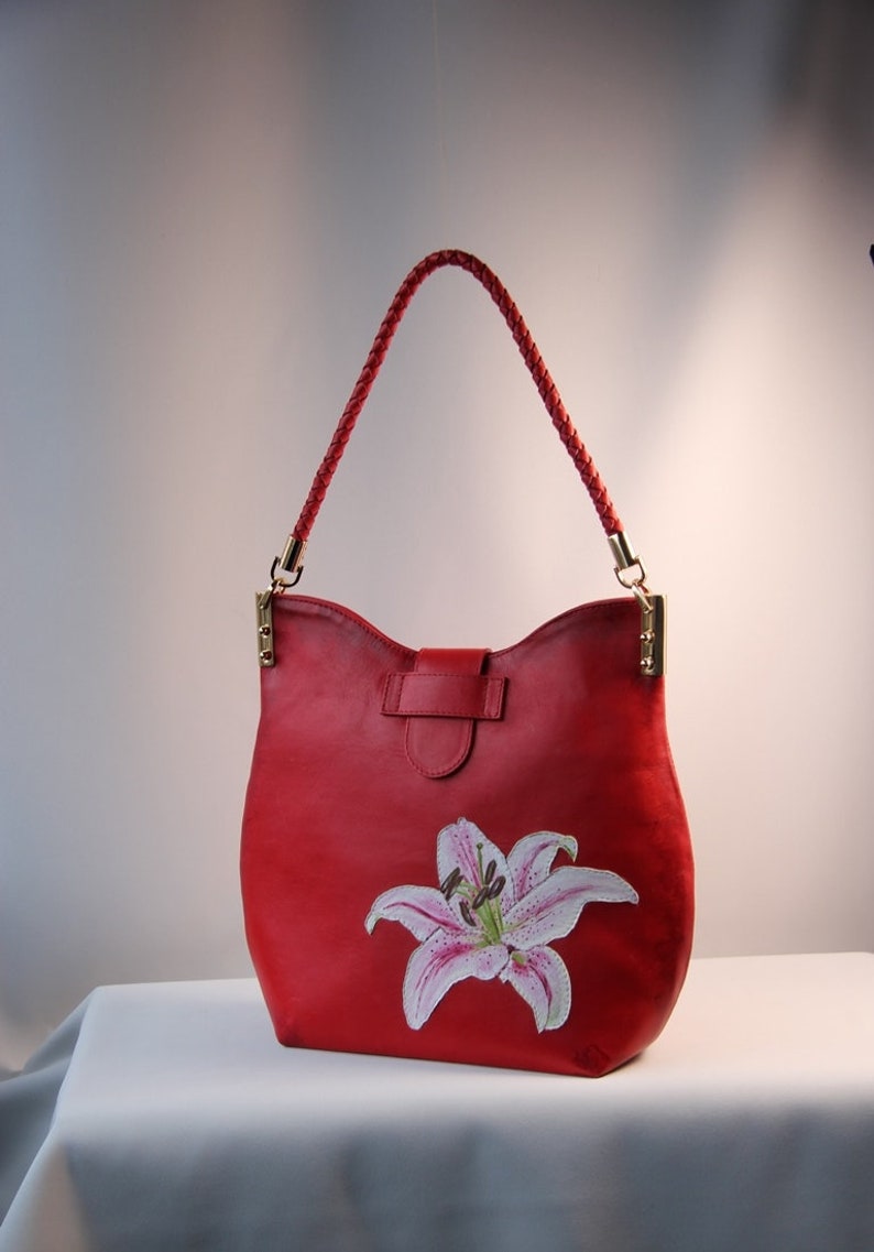 May include: Red leather handbag with a white lily flower painted on the front. The bag has a braided leather strap.
