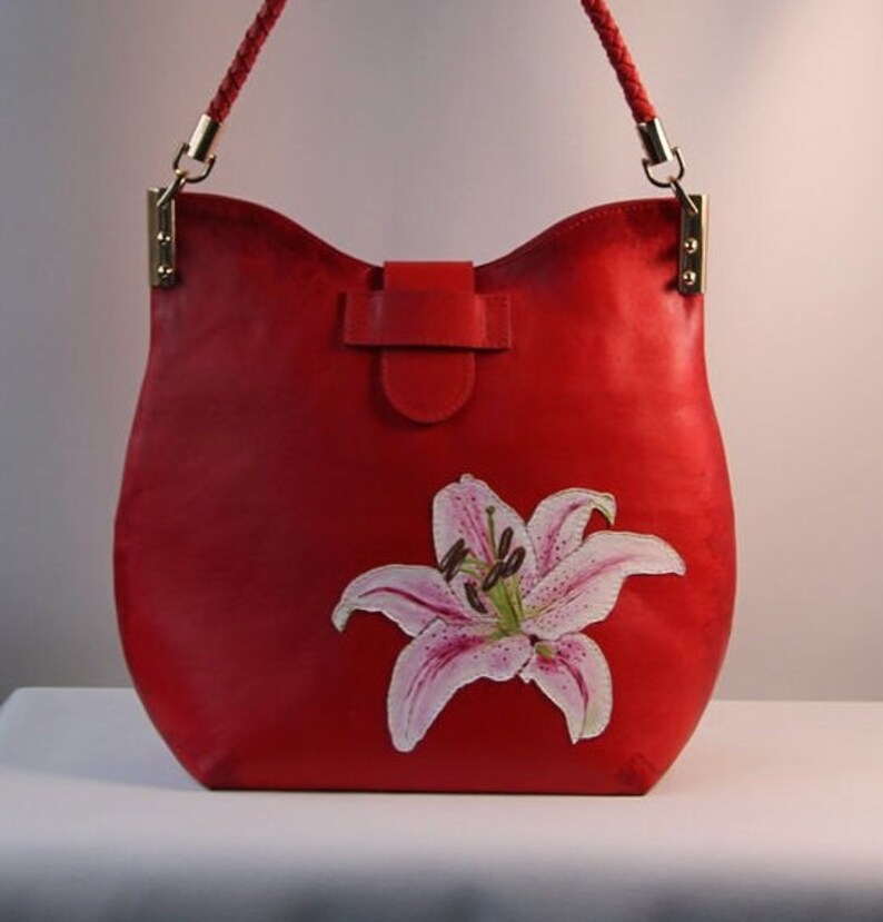 May include: A red leather handbag with a white lily flower design. The bag has a top flap closure and a braided strap.
