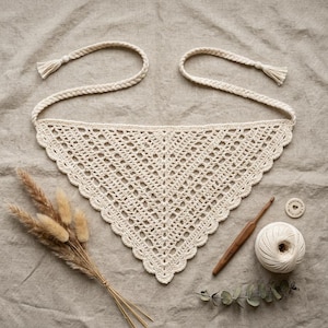 May include: A cream-colored triangular crochet bandana featuring an intricate openwork pattern with a scalloped edge and long braided ties. Included are a wooden crochet hook, a ball of matching yarn, a small wooden tag, and dried grass stems.