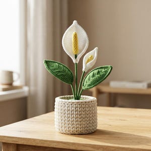 Puede incluir: Una planta de lirio de la paz tejida a crochet con dos espatas blancas, espádices amarillos y dos hojas verdes texturizadas. La planta está en una maceta cilíndrica de crochet color crema con un patrón de puntadas verticales en la base.