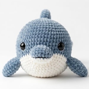 May include: A small, round, crocheted dolphin plush toy featuring a dusty blue upper body and a white underbelly. The toy has two large, glossy black safety eyes, a small dorsal fin on top, and two side fins extending outward from its body.