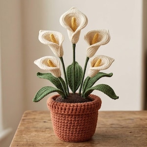 May include: A crocheted potted plant featuring five cream-colored calla lily flowers with yellow spadices, supported by green stems and leaves, all housed in a textured, terracotta-colored crocheted pot with a thick, folded rim.