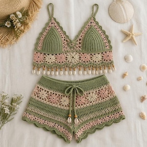May include: A two-piece crochet set consisting of a bralette and shorts. The garments feature a sage green and soft pink floral pattern with scalloped edges. The bralette has thin straps and a row of small white seashells hanging from the hem.