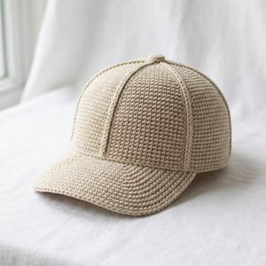 May include: A beige baseball cap constructed from a dense, textured crochet knit. The cap features a structured six-panel design with a curved brim and a small button at the center top, all crafted in a uniform light tan yarn material.