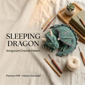 May include: A crocheted green dragon with purple wing accents and cream-colored horns, accompanied by several wooden crochet hooks, a ball of cream yarn, and a small succulent plant. The text reads SLEEPING DRAGON Amigurumi Crochet Pattern.