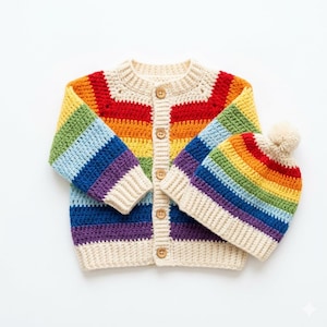 May include: A crochet cardigan and matching beanie set featuring horizontal rainbow stripes in red, orange, yellow, green, blue, and purple. The cardigan has a cream-colored ribbed trim, a round neckline, and five wooden buttons down the front.