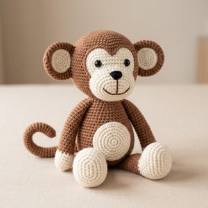 Pode incluir: Um brinquedo de macaco feito em crochê com corpo castanho e rosto, barriga, orelhas e pés na cor creme. O macaco tem olhos de plástico preto, um nariz preto bordado e uma cauda curva. A textura do fio é visível em todo o boneco.
