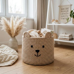May include: A beige knitted storage basket shaped like a bear with two small rounded ears on top. The basket features a stitched black nose and mouth, and two black circular eyes. It is filled with light-colored fabric and wooden baby toys.