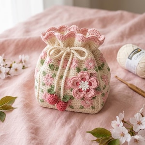 May include: A cream-coloured crocheted drawstring bag, decorated with pink floral embroidery and a large pink flower appliqué. The bag has a ruffled top edge and is tied with a cream-coloured drawstring. Two small pink crocheted berries hang.