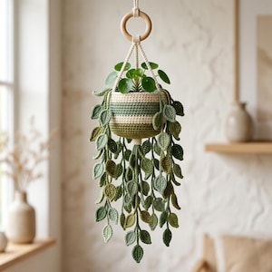 May include: A crocheted hanging planter featuring a cream and green striped basket holding a small green plant. Long, trailing crocheted vines with numerous small green leaves hang down from the basket, suspended by a light-colored wooden ring.