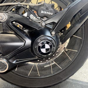 Puede incluir: Un conjunto de rueda trasera de motocicleta en color negro que presenta una tapa de cubo central con el logotipo de BMW. El componente incluye una pinza de freno Brembo negra, un disco de freno de metal perforado y radios metálicos finos.