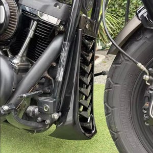 May include: A black motorcycle chin spoiler featuring a V-shaped louvered vent design, mounted on the frame in front of the engine. The component is positioned between the front tyre and the engine block, showcasing a glossy finish and metal hardware.