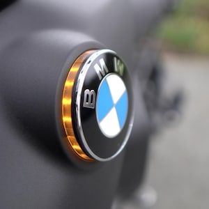 Peut inclure: Gros plan sur un emblème BMW avec un design circulaire bleu et blanc, entouré d'un anneau argenté et d'un fond noir. L'emblème est éclairé par une lumière ambrée.
