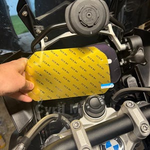 Op de afbeelding: Een gele beschermende schermfolie met een herhalend DCX-9H-patroon en een blauw lipje met het label 2, geplaatst voor het dashboard van een motorfiets. De motorfiets heeft een zwart stuur, een BMW-embleem en een ronde Quad Lock-houder.