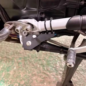 Peut inclure: Un ensemble de béquille latérale de moto comprenant un cadre en métal blanc, un bras de béquille en métal noir et une extension en plastique noir fixée par deux serre-câbles noirs. Le mécanisme inclut des boulons argentés et un ressort.