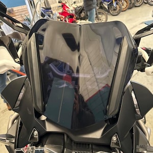 Puede incluir: Primer plano del carenado frontal de una motocicleta, con parabrisas tintado oscuro y componentes negros. El diseño presenta formas angulares y una estética moderna. La imagen está tomada en interiores, con otras motocicletas visibles al fondo.