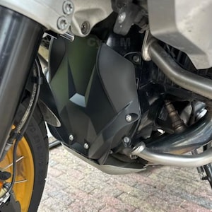 Può includere: Primo piano di un motore di moto con una copertura nera a motivi geometrici. La copertura è fissata con bulloni argentati e si trova sopra un sottoscocca argentato. È visibile una parte del pneumatico anteriore con cerchio dorato.