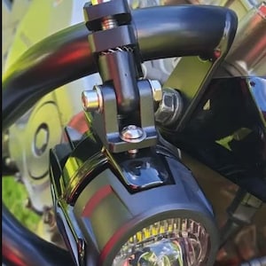Peut inclure: Un phare auxiliaire LED circulaire noir pour moto, fixé sur une barre de protection tubulaire noire à l'aide d'un support métallique. Le phare présente une lentille en verre transparent avec des réflecteurs internes et un boulon argenté.