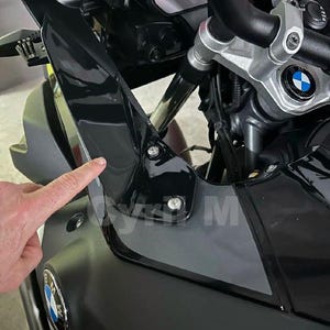 May include: Close-up of a black motorcycle with silver handlebars and a BMW logo. The motorcycle has a black fairing and a digital display. The image is taken from a low angle, showing the front of the bike.