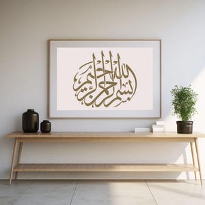 Op de afbeelding: Een ingelijst kunstwerk met gouden Arabische kalligrafie, tentoongesteld op een lichte muur. Het kunstwerk staat boven een houten console met zwarte vazen en een potplant. De kamer heeft een minimalistisch design met natuurlijk licht.