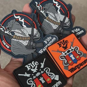 May include: A selection of military-style patches. The patches display a soldier figure with a rifle and smoke, along with the text "Fife Wargames." One patch is orange, and another is black, with additional graphics and text.
