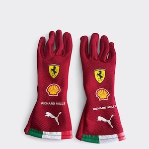 Peut inclure: Une paire de gants de course rouges en tissu maille texturé. Chaque gant présente le logo du cheval cabré Ferrari, un emblème Shell jaune, le texte RICHARD MILLE, un logo Puma blanc et un poignet rayé vert, blanc et rouge.
