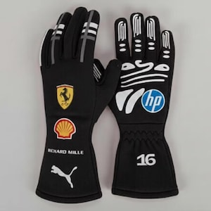 Może przedstawiać: Para czarnych rękawic wyścigowych z białymi i szarymi akcentami. Rękawice posiadają logo Ferrari, logo Shell, tekst Richard Mille i logo Puma. Prawa rękawica ma logo HP i numer 16.