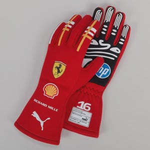 Peut inclure: Une paire de gants de course rouges avec des accents blancs et jaunes. Les gants présentent le logo Ferrari, le logo Shell, le logo Puma et le texte "Richard Mille". Le gant droit porte le numéro "16" et le logo HP.