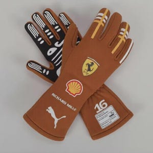 Pode incluir: Dois pares de luvas de corrida cruzadas, uma castanha e outra preta. A luva castanha apresenta riscas brancas e amarelas, um logótipo da Ferrari e um logótipo da Puma. A luva preta tem um design branco. A luva castanha tem o número 16.
