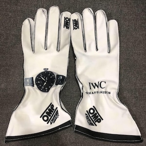 Peut inclure: Une paire de gants de course blancs avec des bordures noires. Le gant gauche présente un graphique de montre-bracelet à cadran noir. Le gant droit affiche le texte IWC SCHAFFHAUSEN. Les deux gants portent le logo OMP en noir sur les poignets.