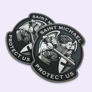 Puede incluir: Dos parches de San Miguel en negro y gris. Cada parche presenta una ilustración de San Miguel con una espada y un escudo, rodeada de las palabras "Saint Michael" y "Protect Us".